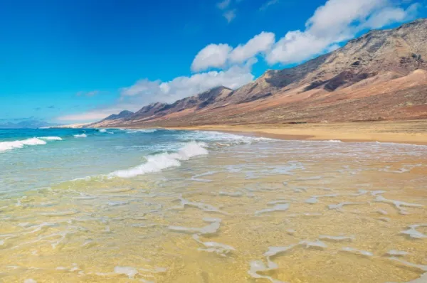 Spanische Insel Fuerteventura: idyllischer Strand mit großen Dünen im Hintergrund bei klarem Himmel.