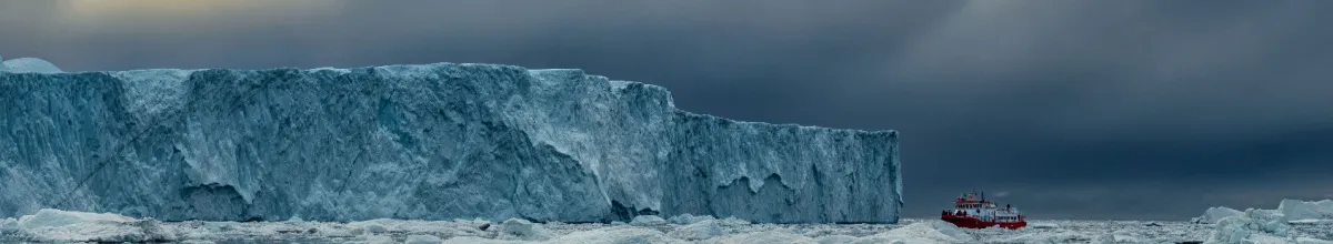 Eine riesige Eismasse am Meer und ein Schiff, welches sie umschifft an einem wolkenverhangenen Tag.