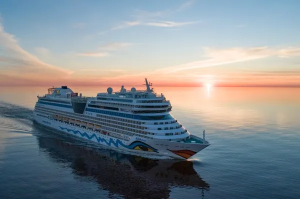 AIDAmar von seitlich vorne im Sonnenuntergang auf dem Meer. Im Hintergrund ein blauer Himmel mit rötlich schimmerndem Horizont und einzelnen weißen & rötlichen Cirrus-Wolken