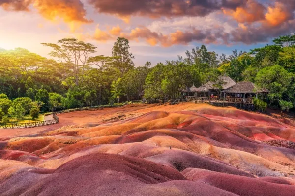 Siebenfarbige Erde auf Madagaskar Die siebenfarbige Erde - bestehend aus Aluminium- und Eisenoxiden - bei Charmarel auf Madagaskar. Im Hintergrund einige Bäume und ein Holzhaus, darüber ein leicht bewölkter Himmel im goldenen Licht des Sonnenuntergangs.