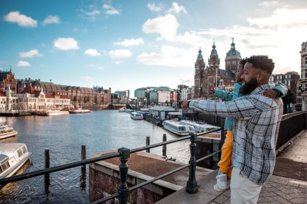 NCL Free at Sea – Abenteuer für die ganze Familie im Hafen Ein Vater hält sein Kind im Arm und deutet auf die Grachten und historischen Gebäude von Amsterdam während einer Stadtbesichtigung auf einer Kreuzfahrt mit Norwegian Cruise Line.