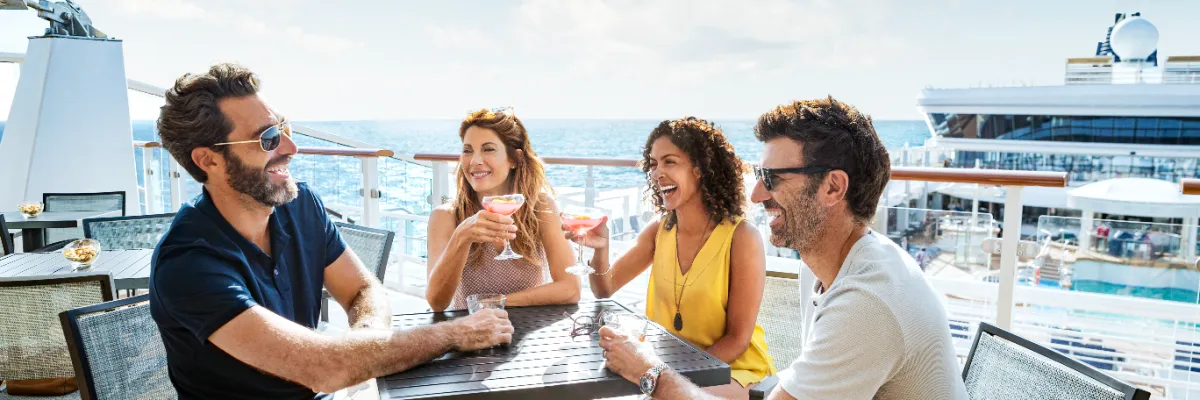 NCL Open Bar – Gesellige Stunden auf dem Außendeck genießen Vier gut gelaunte Gäste sitzen an einem Tisch auf dem Sonnendeck eines NCL-Kreuzfahrtschiffs, unterhalten sich angeregt und stoßen mit farbenfrohen Cocktails an.