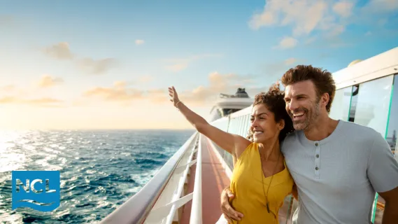 Ein lächelndes Paar steht Arm in Arm an der Reling eines Norwegian Cruise Line Kreuzfahrtschiffs und blickt bei Sonnenuntergang auf das weite Meer.