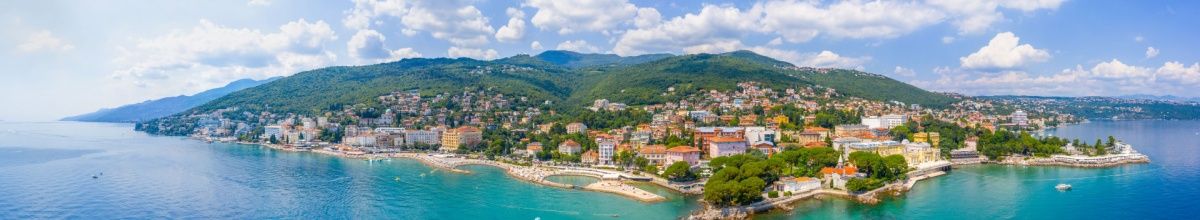 Weitwinkelperspektive auf die Stadt Opatija mit kleinen Häusern im Vordergrund und einer grünen Berglandschaft dahinter.