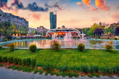 Das Stadtbild von Tirana Ein Einblick in die Stadt Tirana bei Abenddämmerung mit Aussicht auf einen Park mitsamt Springbrunnen und Hausfassaden im Hintergrund.