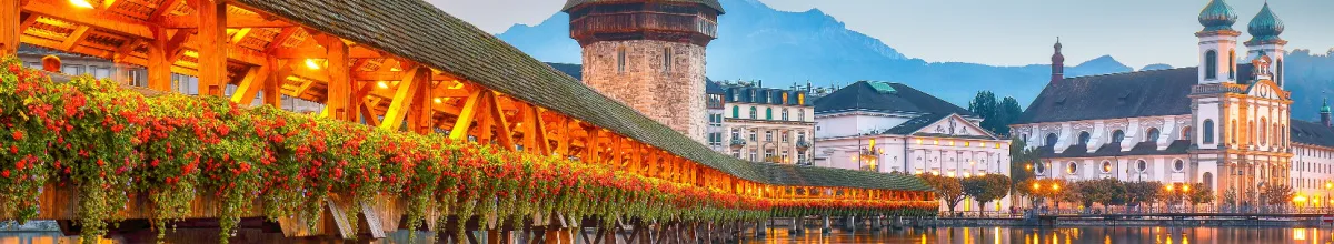 Eine warm beleuchtete altertümliche Brücke mit Dach, die ins Zentrum von Luzern führt mit einem Wachturm und der Kulisse der Stadt im Hintergrund bei Tag.