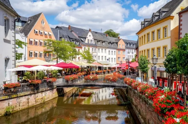 Saarburg Ein Flusslauf fließt durch die Stadt Saarburg umgeben von schönen kleinen Häusern in der Sonne und roten Blumen am Geländer.