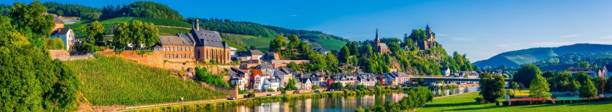 Blick auf die Stadt Saarburg mit kleinen, bunten Häusern am Ufer und Kirche und Ruine auf einem Hügel im Hintergrund.
