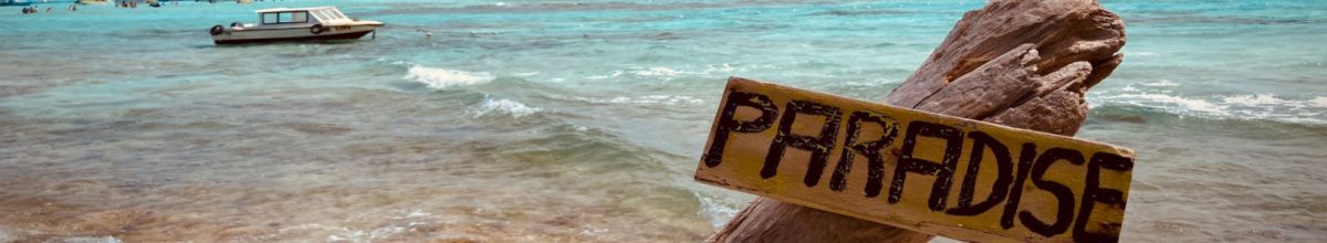 Ein krudes Holzschild am Strand von Providencia mit der Aufschrift "Paradise" und einem kleinen Boot im Hintergrund bei Tag.