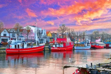Ein Ufer innerhalb der Stadt mit einigen rot-weißen Fischerbooten und den bunten Häusern der Stadt im Hintergrund bei einem malerischen Sonnenuntergang.