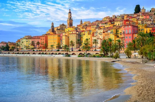 Die farbenprächtige Altstadt von Menton, mit gelben und pinken Häusern, Palmen und einem Sandstrand im Vordergrund.