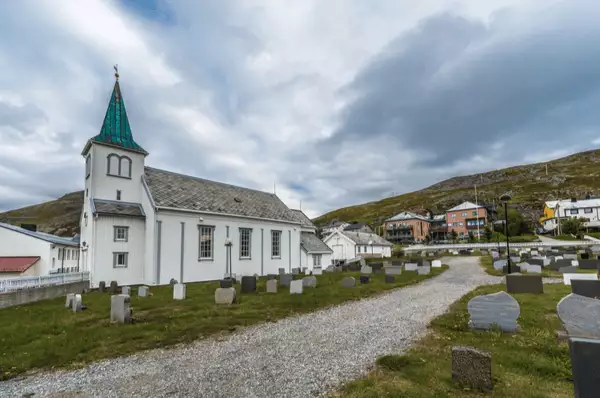 Seitenansicht der weißen Kirche von Honningsvåg. Davor der Friedhof und ein Gehweg, im Hintergrund die Häuser des Ortes.