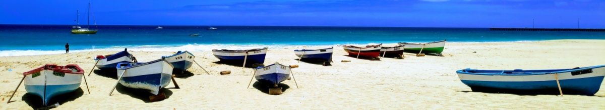 Kleine Fischerboote im weißen Sand vor türkisfarbenem Meer unter blauem Himmel.