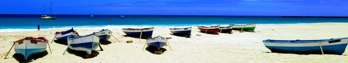 Kleine Fischerboote im weißen Sand vor türkisfarbenem Meer unter blauem Himmel.