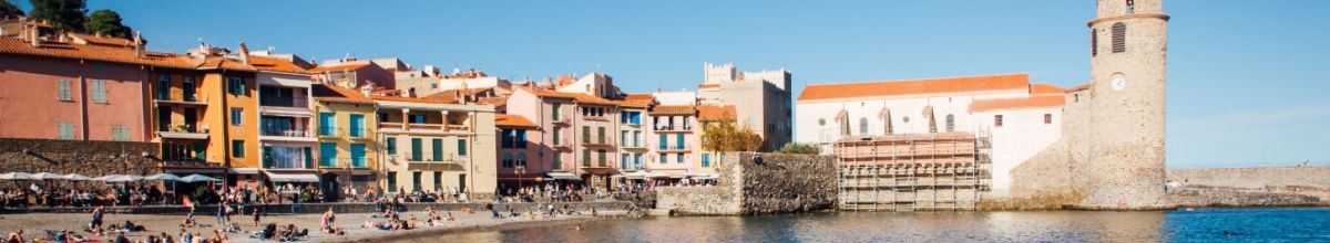 Die Hafenkante von Collioure bei Tag, mit einer alten Befestigungsanlage, Strand und den Häusern der Stadt.