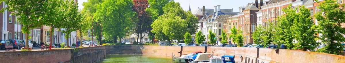 Eine Gracht in Middelburg mit Booten vor Anker, baumgesäumten Alleen nebendran und den Häusern der Stadt bei Tag.