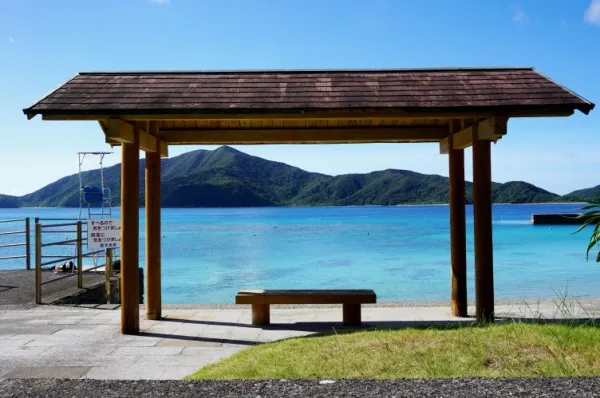 Ein Unterstand mit einer Bank am Tag mit Ausblick auf das azurblaue Meer und grünen Bergen im Hintergrund.