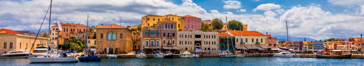 Eine bunte, sonnenbeschienene Häuserfront in Chania mit einigen angetauten Booten davor.