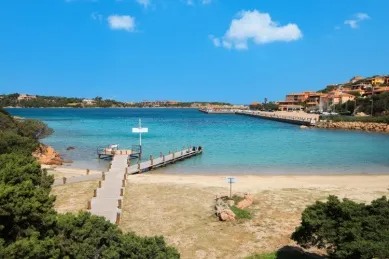 Der Strand bei Porto Cervo Ein Sandstrand bei Porto Cervo am Tag mit einem Kai und der Stadt rechts.