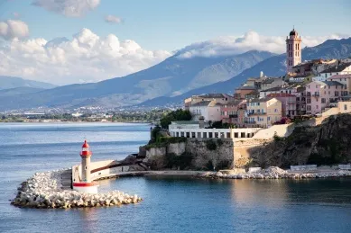 Bastia Hafeneingang von Bastia mit kleinem roten Leuchtturm in Vordergrund und der Stadt und Bergen im Hintergrund.