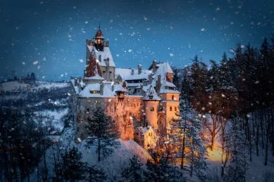 Ein großes mittelalterliches Schloss bei Nacht inmitten eines Schneegestöbers kontrastiert durch warmes Licht am Eingang.
