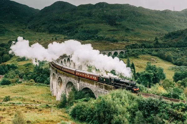 Der Hogwarts Express, eine alte Dampflock, fährt durch eine bergige Landschaft auf einer Brücke.