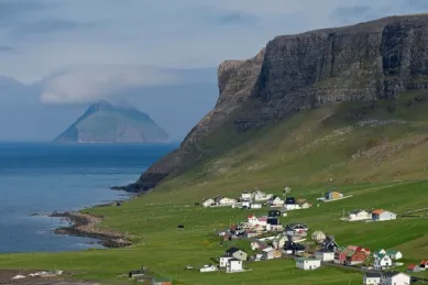 Ein Teil der Küste Suðuroys und eine kleine Ortschaft inmitten einer wallenden, grünen Wiese mit einem Bergplateau im Hintergrund bei Tag.