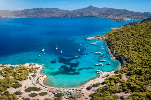 Ägina Strand Eine Bucht mit türkisfarbenem Meer und einigen Booten, die vor Land ihren Anker gesetzt haben, drumherum grüne Natur.