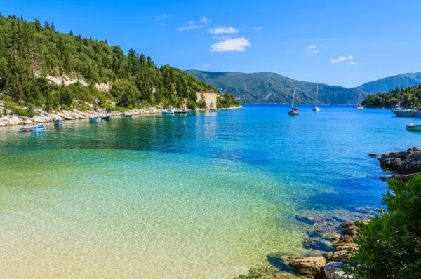 Fiskardo Strand Strand mit ruhigem, klarem Wasser umgeben von grüner Natur, auf dem Wasser sind einzelne kleine Boote zu sehen.
