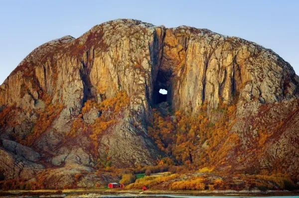 Imposant ragt der Fels Torghatten mit seinem Loch empor. An seinem Fuße sind kleine rote Häuser sichtbar.