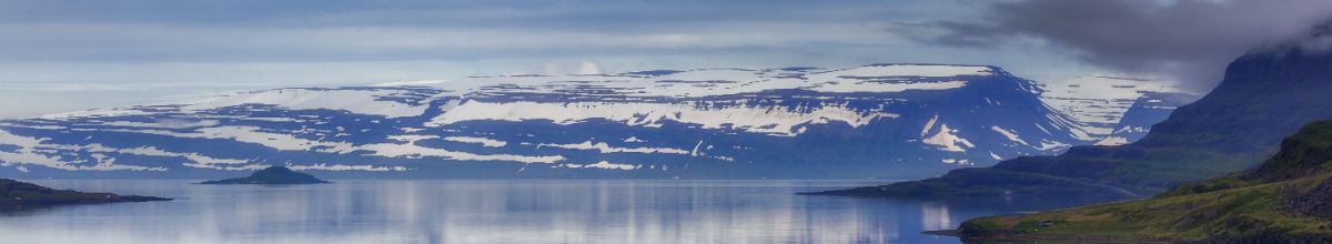 Die eisige Bergkulisse Islands von der Insel Vigur aus betrachtet mit einem Meerarm im Vordergrund.