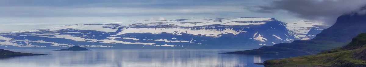 Die eisige Bergkulisse Islands von der Insel Vigur aus betrachtet mit einem Meerarm im Vordergrund.
