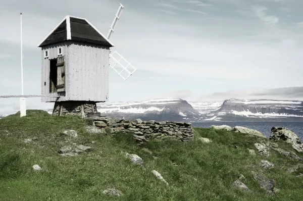 Eine Holzwindmühle auf Vigur Eine alte Holzwindmühle auf Vigur mit einem kleinen steinernen Wall und dem riesigen Bergmassiven Islands im fernen Hintergrund bei Tag.
