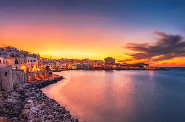 Das pittoreske Stadtbild von Vieste am Abend mit den warmen Lichtern der Stadt und dem ruhigen, angrenzenden Mittelmeer von einem nahen Steinstrand aus fotografiert.