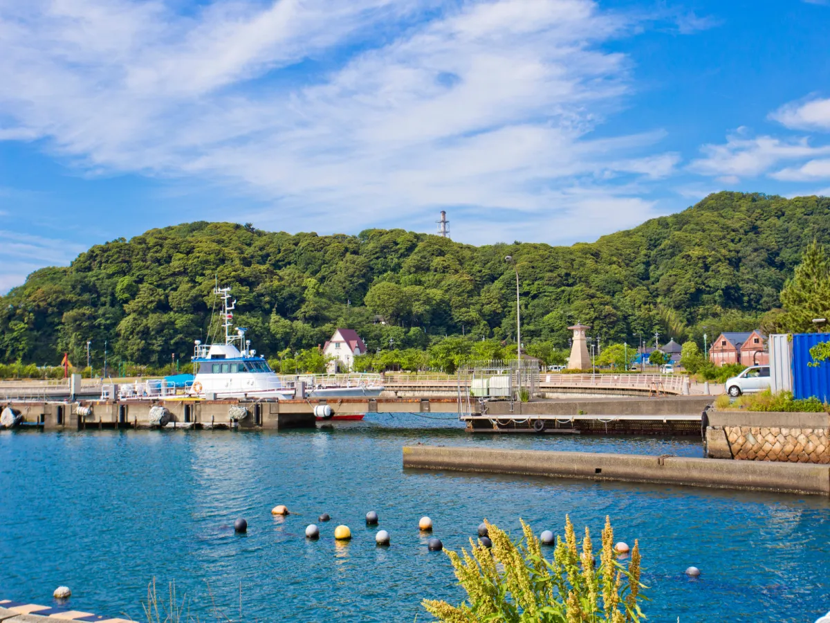 Der Port von Tsuruga am Tag mit klarem, blauen Wasser, bewaldeten Hügeln und den Hafengebäuden.