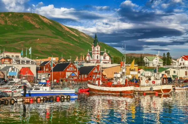 Island Kreuzfahrt Hafen – Traditionelles Küstenflair und bunte Fischerboote Bunte traditionelle Isländer-Holzhäuser und Holz-Fischerboote im ruhigen Hafenbecken vor einer malerischen Bergkulisse und einer typisch isländischen Holzkirche.