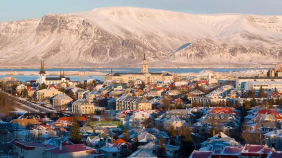 Panoramaansicht der verschneiten bunten Häuser von Reykjavík vor dem im Sonnenlicht erstrahlenden Hausberg Esja.