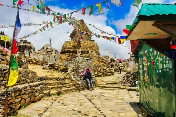 Mongla Eine Wanderin sitzt auf Steinen im Ort Mongla, darüber hängen bunte Girlanden unter blauem Himmel.