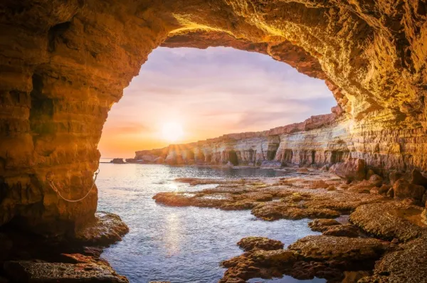 Beste Reisezeit nach Zypern Eine Grotte mit orangenem Gestein. Im Hintergrund scheint die Sonne über das Meer.