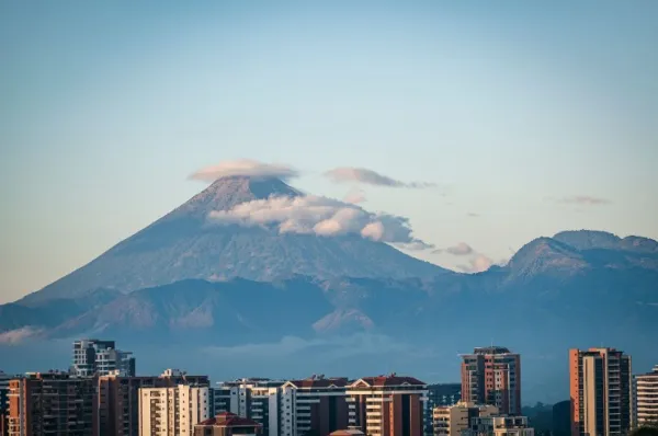 Hochhäuser stehen vor dem Panorama eines wolkenverhangenen Vulkans in Guatemala.