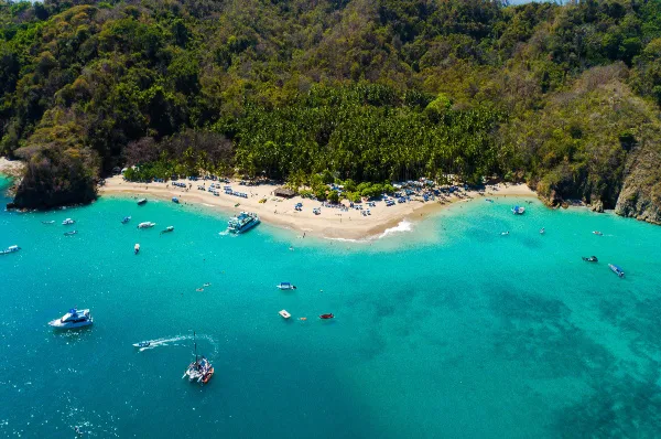 Die Küste der Isla Tortuga mit vielen Schiffen im türkisen Wasser vor Anker, einem kleinen Sandstrand und dem Regenwald der Insel bei Tag.