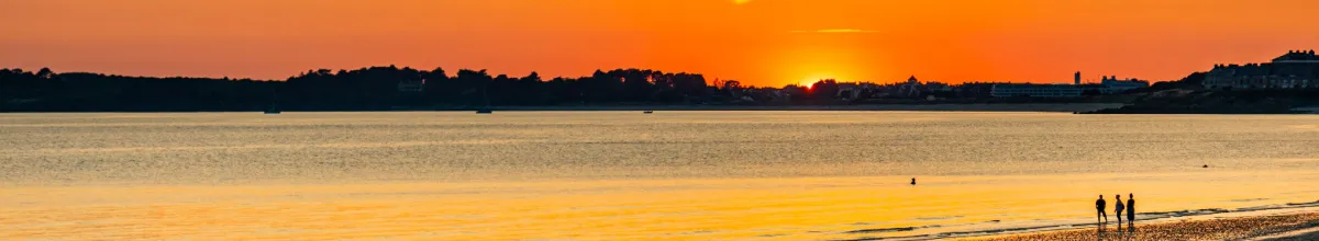 Ein malerischer Sonnenuntergang über der Hafenstadt Quiberon mit dem Meer im Vordergrund und der Silhoutten der Stadt in der Ferne.