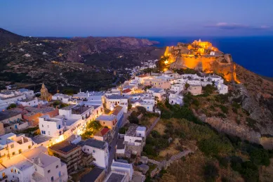 Die Stadt Kythira fotografiert von einem nahen Hügel bei Nacht, ausgezeichnet durch weiße Hausfassaden, warme Lichter und ein altes Kastell, das über der Stadt thront.