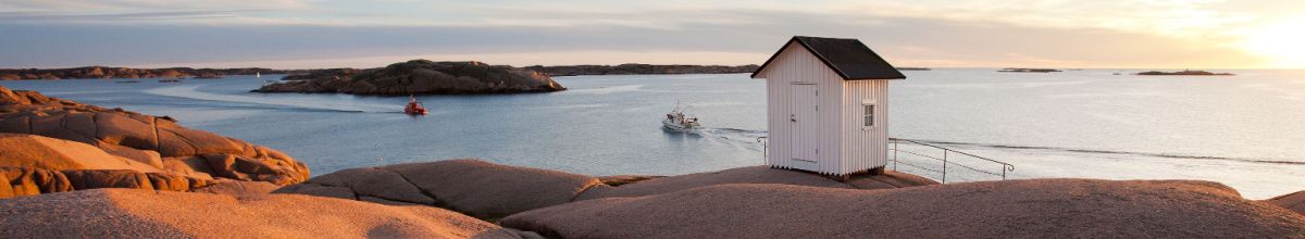 Ein kleines, weißes Haus auf einem Felsen an der Küste von Lysekil bei Sonnenuntergang, im Hintergrund ist das Meer mit zwei Schiffen.