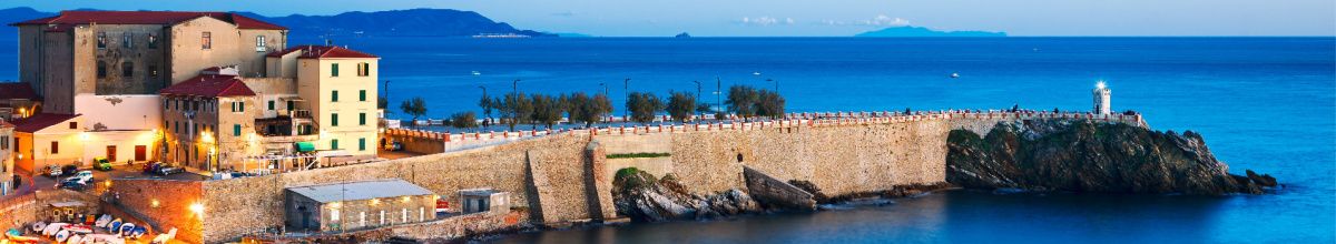 Die steinerne Hafenmauer von Piombino am Abend, warm beleuchtet mitsamt einer schnuckeligen Promenade.