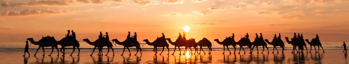 Eine Reihe Kamele reitet beim Sonnenuntergang entlang des Strandes bei Broome in Australien.