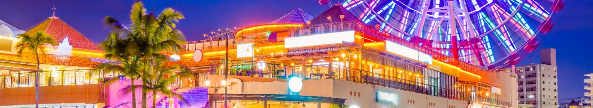 Teil einer Vergnügungsmeile in Okinawa mit hell beuchteten Gebäuden am Abend und einem Riesenrad im Hintergrund.