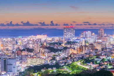 Die bunt und hell erleuchtete Sykline der Stadt Okinawa bei Sonnenuntergang, geprägt durch viele Hochhäuser und einen unverkennbaren, urbanen Charme.