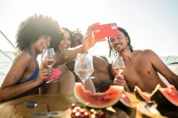 Genuss in Vollpension - auch im Young & Young Tarif von MSC Vier junge Leute genießen Wein und Wassermelonen an Bord eines Kreuzfahrtschiffes und machen Selfies dabei.