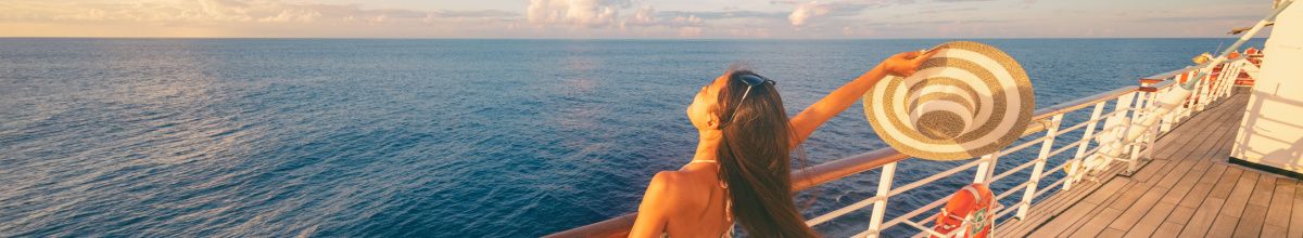 Frau mit Sonnenhut an der Reling eines Kreuzfahrtschiffes blickt bei Sonnenuntergang auf das offene Meer.